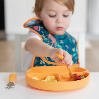 A child in a jungle-themed bib eats from an orange plate with matching utensils, all from Bumkins Ultimate Gift Set - Jungle & Animal Prints.