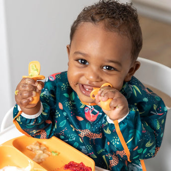 A baby in a high chair eats from a plate with utensils, wearing a jungle-themed bib from Bumkins Ultimate Gift Set - Jungle & Animal Prints.