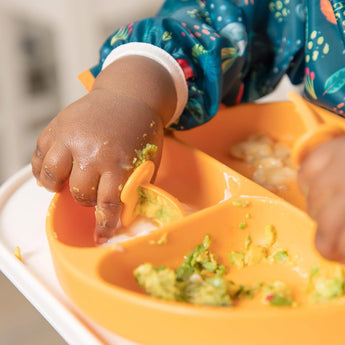 Close-up of a child's hand in a sleeved bib eating from a plate with utensils, all from Bumkins Ultimate Gift Set - Jungle & Animal Prints.