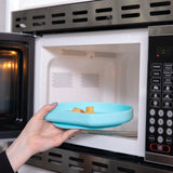A hand places a Bumkins Silicone Grip Plate: Blue filled with food into an open microwave.