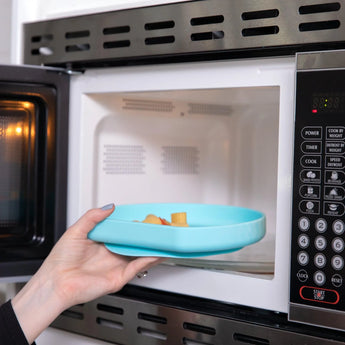 A hand places a Bumkins Silicone Grip Plate: Blue filled with food into an open microwave.