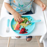 A child in a dinosaur-print bib seated in a high chair eats peas, watermelon, and pasta from a Bumkins Silicone Grip Plate: Blue.