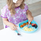 A child in a purple bib enjoys eggs, potatoes, and fruit from a Bumkins Silicone Grip Plate: Blue with a fork on a white table.