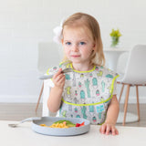 A toddler with a bow happily eats macaroni and strawberries from a Bumkins Silicone Grip Plate: Gray at a white table, wearing a cactus bib.