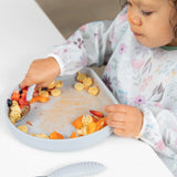 A toddler in a floral bib enjoys pasta and veggies with a spoon from a Bumkins Silicone Grip Plate: Gray on a white table.