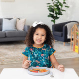 A smiling girl with curly hair enjoys chicken nuggets from a Bumkins Silicone Grip Plate: Gray at a white table in a cozy living room.
