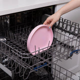 A hand places a Bumkins Silicone Grip Plate: Pink into the top rack of an open dishwasher.