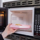 A hand places a Bumkins Silicone Grip Plate: Pink filled with food into an open microwave.