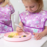Two toddlers in purple bibs sit at a white table. One uses a spoon to eat scrambled eggs from a Bumkins Silicone Grip Plate: Pink.