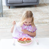 A young girl in a purple bib removes a lid from a Bumkins Silicone Grip Plate: Pink with a sandwich & strawberries on a white table.
