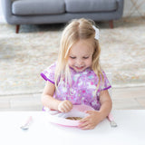 A girl in a purple bib smiles at a white table, removing a lid from a Bumkins Silicone Grip Plate: Pink while a spoon and fork rest nearby.