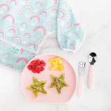 Bumkins Silicone Grip Plate: Pink with star-shaped toast and fruit sits next to utensils and a rainbow-print bib on a marble surface.