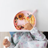 Top-down view of a child enjoying fruit, eggs, and toast from a Bumkins Silicone Grip Plate: Pink, wearing a floral bib at a white table.