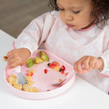 A toddler enjoys pasta, avocado, and strawberries with a fork & spoon from a Bumkins Silicone Grip Plate: Pink on a white table.