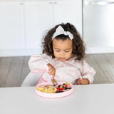 A toddler enjoys scrambled eggs and berries from a Bumkins Silicone Grip Plate: Pink while sitting in a white kitchen.