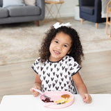 A young girl smiles, wearing a heart-patterned bib while holding a fork over a Bumkins Silicone Grip Plate: Pink on a white table.