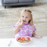 A young girl enjoys sliced fruits and toast from a Bumkins Silicone Grip Plate: Pink, wearing a purple bib with cute designs.