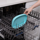 A hand places a Bumkins Silicone Grip Tray: Blue into the top rack of an open dishwasher.