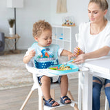 A toddler wearing a bib in a high chair enjoys noodles from a Bumkins Silicone Grip Tray: Blue, as a woman nearby holds a fork of food.