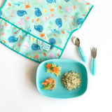 Bumkins Silicone Grip Tray: Blue with quinoa and veggies sits near an ocean-themed bib and blue utensils on a white background.