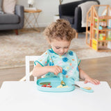 A toddler with curly hair eats from a Bumkins Silicone Grip Tray: Blue at a white table, with toys and a sofa in the background.