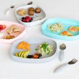 Four plates, including Bumkins Silicone Grip Tray: Gray, are arranged with various foods and utensils on a white background.