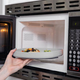 A hand places a Bumkins Silicone Grip Tray: Gray filled with food into an open microwave.