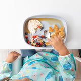 Top-down view of a child enjoying pancakes with fruit and yogurt on a Bumkins Silicone Grip Tray: Gray, wearing a sea creature bib.