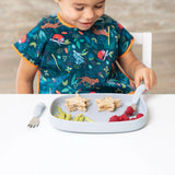 A child in a bib sits at a table, eating star sandwiches, green snacks, and raspberries from Bumkins Silicone Grip Tray: Gray using cutlery.
