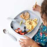A child in a colorful bib enjoys eggs, fruit, and oats from a Bumkins Silicone Grip Tray: Gray at a white table with utensils nearby.