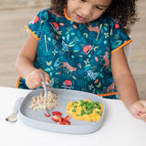 A child in a colorful bib enjoys chicken, strawberries, and macaroni with peas from a Bumkins Silicone Grip Tray: Gray at a white table.