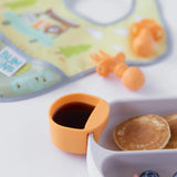 Close-up of a Bumkins Silicone Little Dipper 3-Pack: Tutti-Frutti with syrup, with a plate, utensils and bib nearby on a white background.