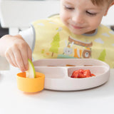 A child in a bib smiles while dipping an apple slice in an orange Bumkins Silicone Little Dipper 3-Pack: Tutti-Frutti attached to a plate.