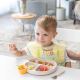 A toddler smiles at a table, enjoying snacks with a fork from a plate with an orange Bumkins Silicone Little Dipper 3-Pack: Tutti-Frutti.