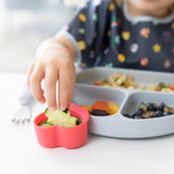 Close-up of a child joyfully dipping a cucumber in a red Bumkins Silicone Little Dipper 3-Pack: Tutti-Frutti attached to a plate of food.