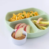 A green plate with food, and a marble Bumkins Silicone Little Dipper Round 3-Pack: Taffy with peanut butter on a white background.