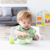 A curly-haired toddler holds a spoon near a green bowl with a green Bumkins Silicone Little Dipper Round 3-Pack: Taffy attached.