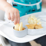 Close-up of a child dipping apple slices into peanut butter in a gray Bumkins Silicone Little Dipper Round 3-Pack: Taffy attached to a plate.