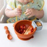 A child in a high chair, enjoying fruit from a bowl with a gray Bumkins Silicone Little Dipper Round 3-Pack: Taffy with peanut butter.