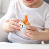 A child in a white shirt holds a marble Bumkins Silicone Little Dipper Round 3-Pack: Taffy filled with carrot sticks.