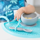 Close-up of a baby reaching for a gray bowl of food on a Bumkins Silicone Sensory Placemat: Blue, with a gray spoon attached to a tether.