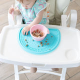 A baby uses a pink spoon and bowl on a Bumkins Silicone Sensory Placemat: Blue on a white high chair tray.