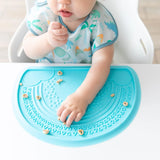 A baby eagerly eats cereal on a Bumkins Silicone Sensory Placemat: Blue, wearing a dinosaur-patterned bib in a high chair.