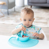 A toddler in a bib happily eats cereal from a blue bowl on a Bumkins Silicone Sensory Placemat: Blue.