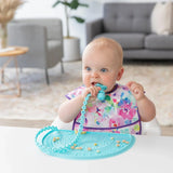 A baby chews a blue silicone utensil near a Bumkins Silicone Sensory Placemat: Blue with snacks scattered, in a cozy living room.
