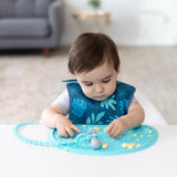 A toddler playfully explores food on a Bumkins Silicone Sensory Placemat: Blue in a bright living room.