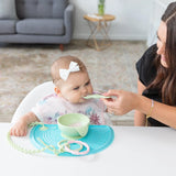 A woman feeds a baby wearing a headband and bib from a green bowl on a Bumkins Silicone Sensory Placemat: Blue.