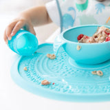 Close-up of a child in a bib spilling water from a blue cup on a Bumkins Silicone Sensory Placemat: Blue near a blue bowl with oatmeal.