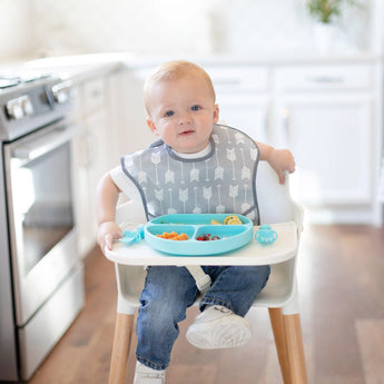 A baby in a high chair wears a Bumkins SuperBib®: Arrow, enjoying snacks from a blue plate in a bright kitchen.