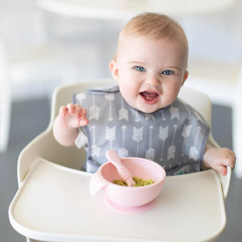 A baby in a high chair, wearing a Bumkins SuperBib®: Arrow, eats mashed avocado from a pink bowl with a matching spoon.